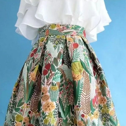 Weiße Bluse mit floralem, buntem Faltenrock vor blauem Hintergrund.