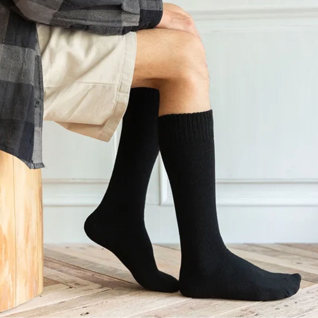 Mann trägt schwarze Kniestrümpfe aus Baumwolle, sitzt auf Holzbank, beige Shorts.