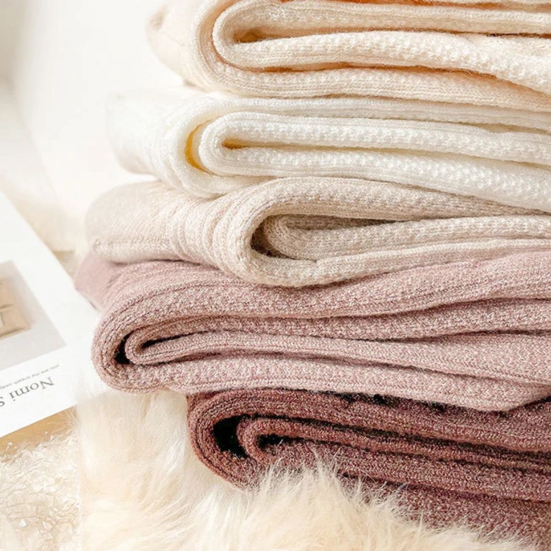 Gestapelte, weiche, beige und rosa Strickdecken aus Baumwolle auf einem flauschigen Untergrund.