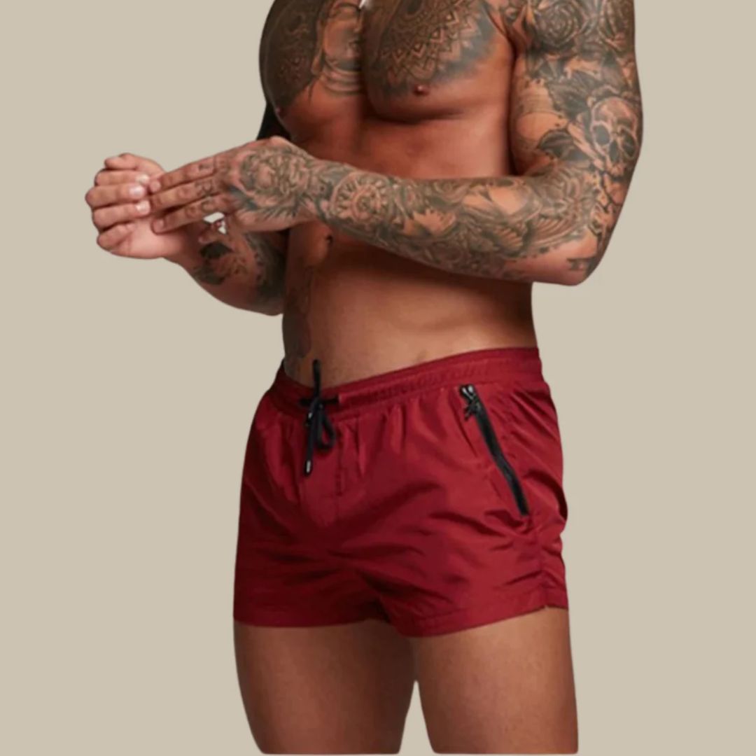 Mann mit tätowiertem Oberkörper trägt rote Badehose, sportlich, Sommermode, Herrenbekleidung.