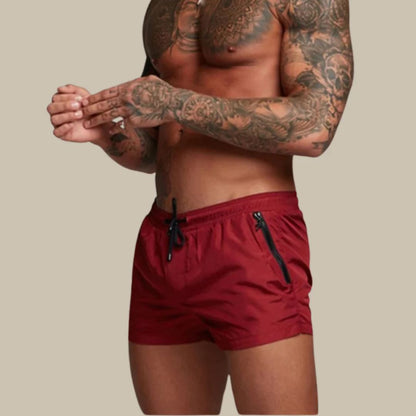 Mann mit tätowiertem Oberkörper trägt rote Badehose, sportlich, Sommermode, Herrenbekleidung.