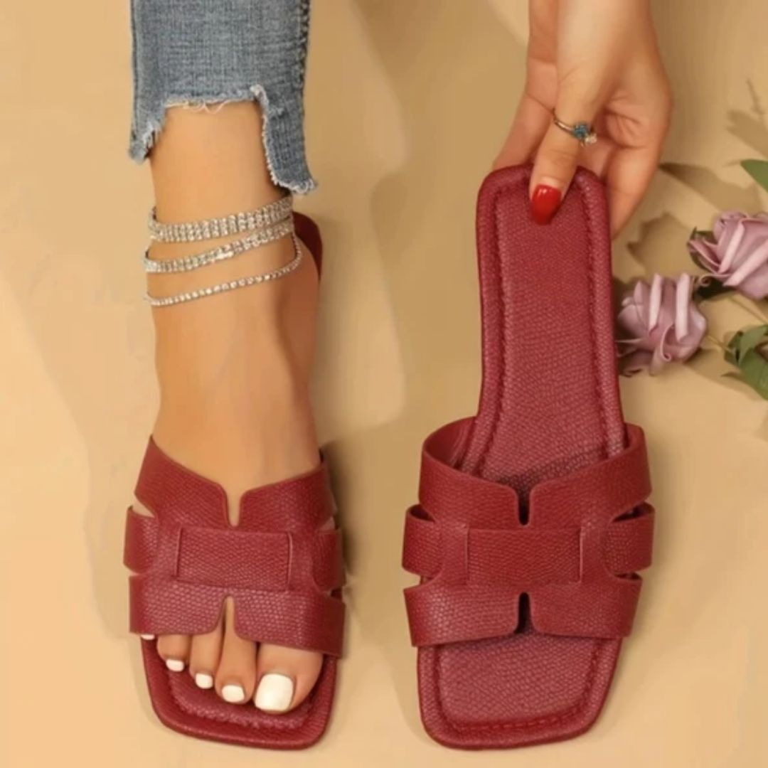 Rote Damen-Sandalen aus Leder, flach, modisch, mit Fußkettchen, neben Rosen.