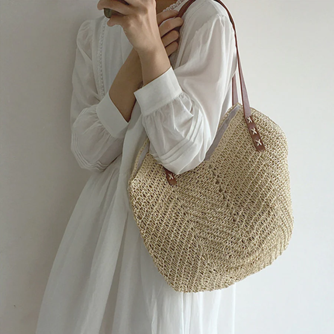 Frau mit weißem Kleid trägt beige Strohtasche mit braunen Riemen.