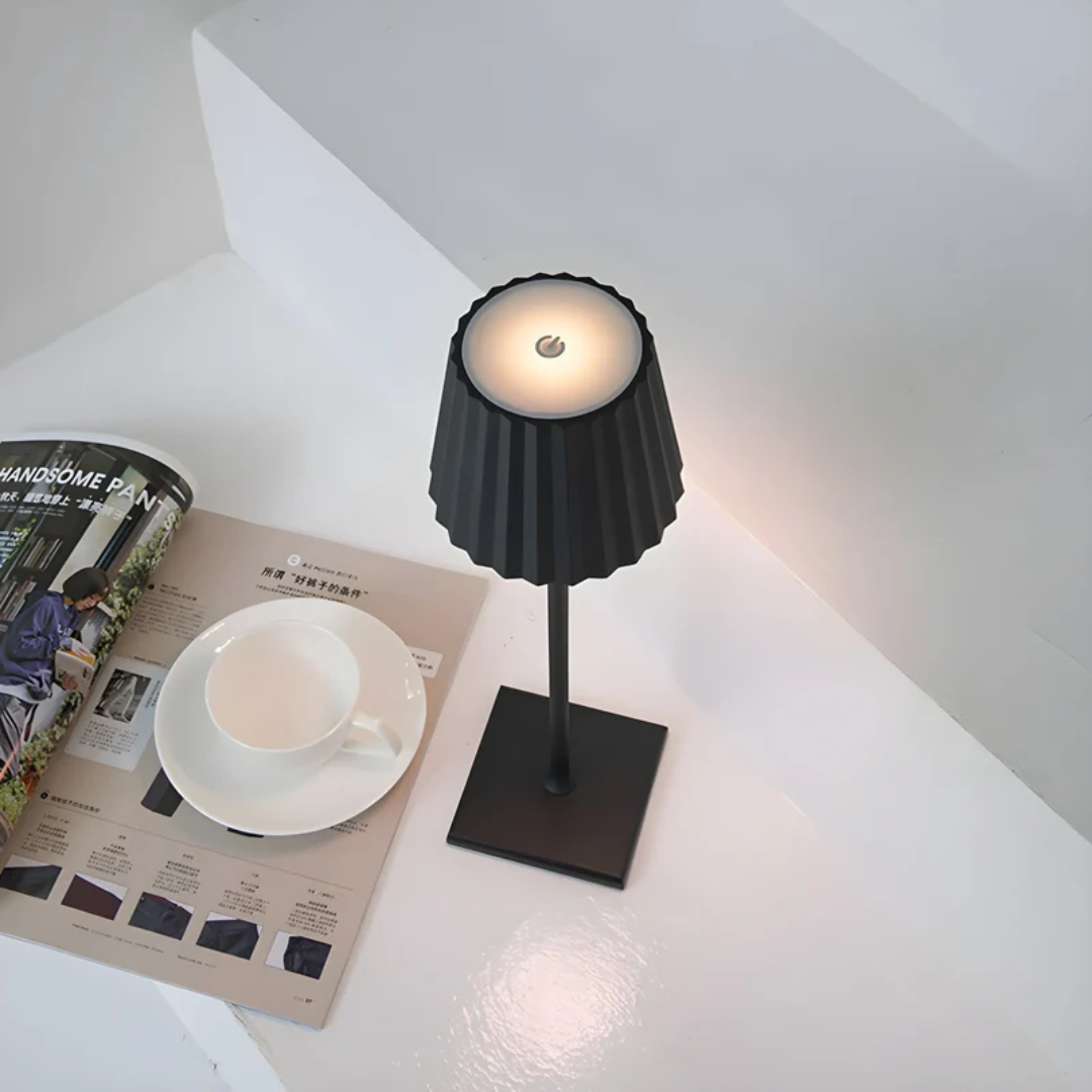 Schwarze Tischlampe mit geriffeltem Schirm auf weißem Tisch neben Tasse und Magazin.