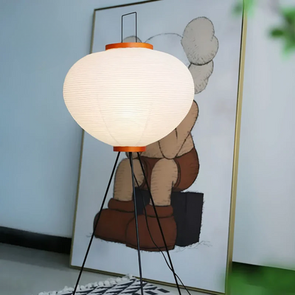 Moderne Stehlampe mit weißem Lampenschirm und schwarzem Stativ, vor Kunstwerk im Hintergrund.