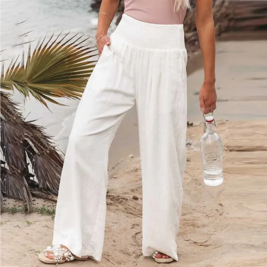 Frau in weißen Leinenhosen am Strand, hält Wasserflasche, Sommermode, lässiger Look.