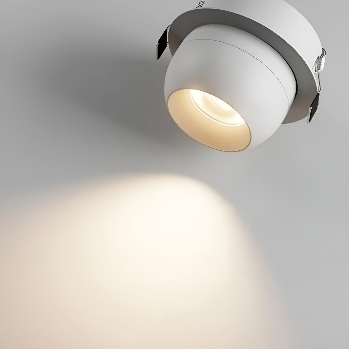 Närbild underifrån på en vit infälld spotlight med klämfjädrar och en mjuk, varm ljuskägla på väggen 4