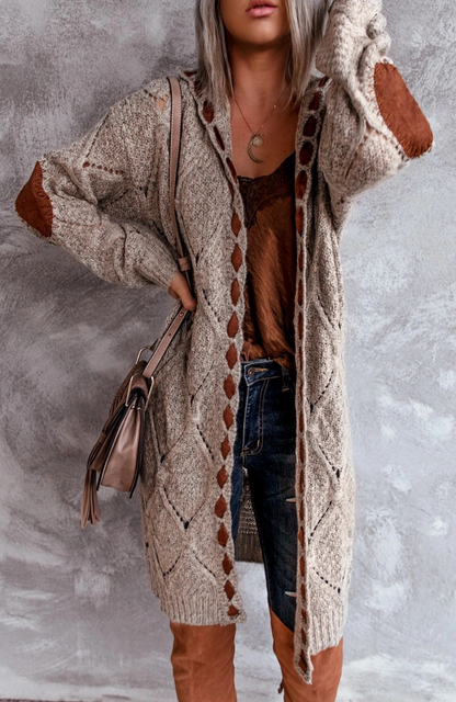 Frau in beigem Strickcardigan mit braunen Patches, Jeans und braunen Stiefeln vor grauem Hintergrund.