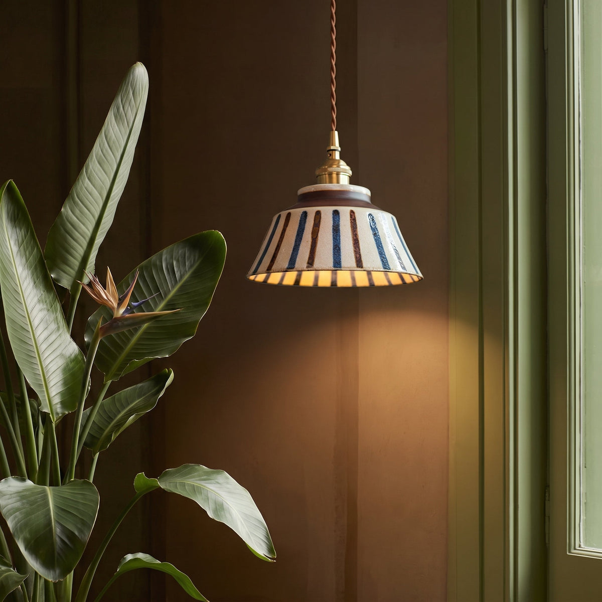 Tänd lampa med randig keramikskärm och mässingsfattning hänger bredvid en stor växt med blomma mot en mörkbrun vägg. 4