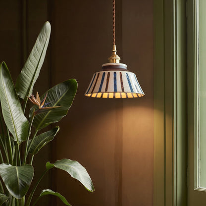 Tänd lampa med randig keramikskärm och mässingsfattning hänger bredvid en stor växt med blomma mot en mörkbrun vägg. 4