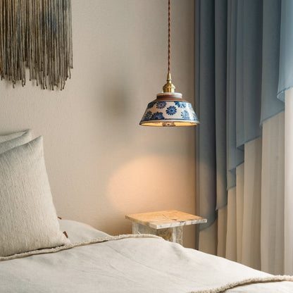 Tänd lampa med blå blommönstrad keramikskärm hänger vid en säng med beige textilier, ett marmorbord och blå gardiner. 2