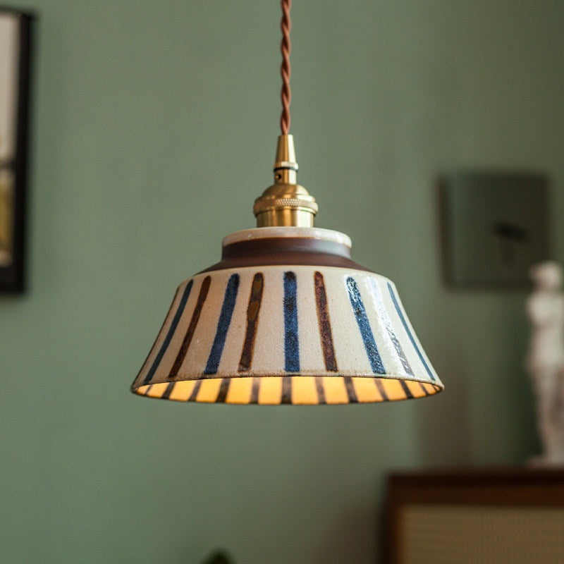 Lampa med randig keramikskärm i blått och brunt och flätad sladd hänger tänd mot en grön vägg. 3
