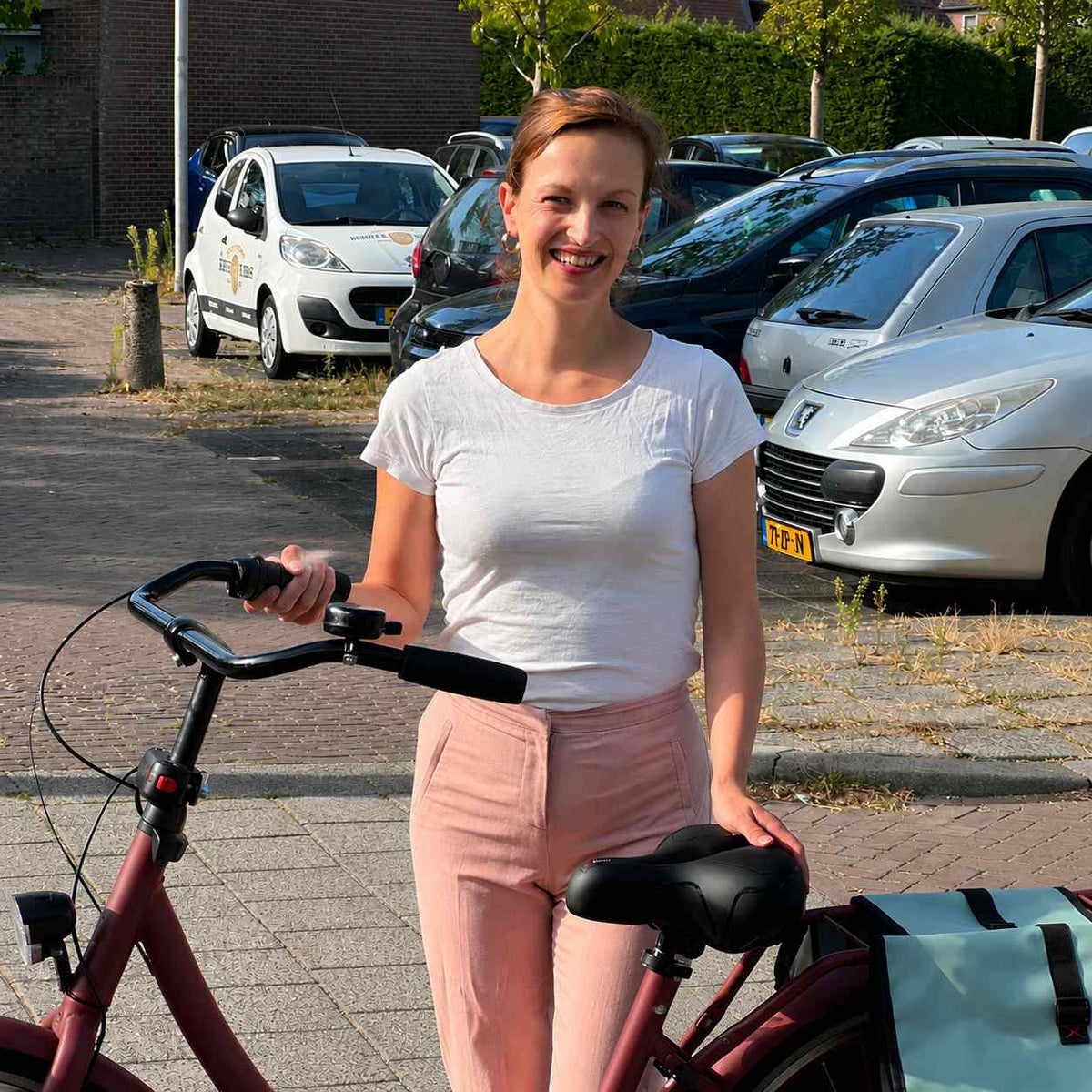 Leende kvinna står bredvid sin röda cykel och lägger handen på den svarta sadeln på en parkering. 7