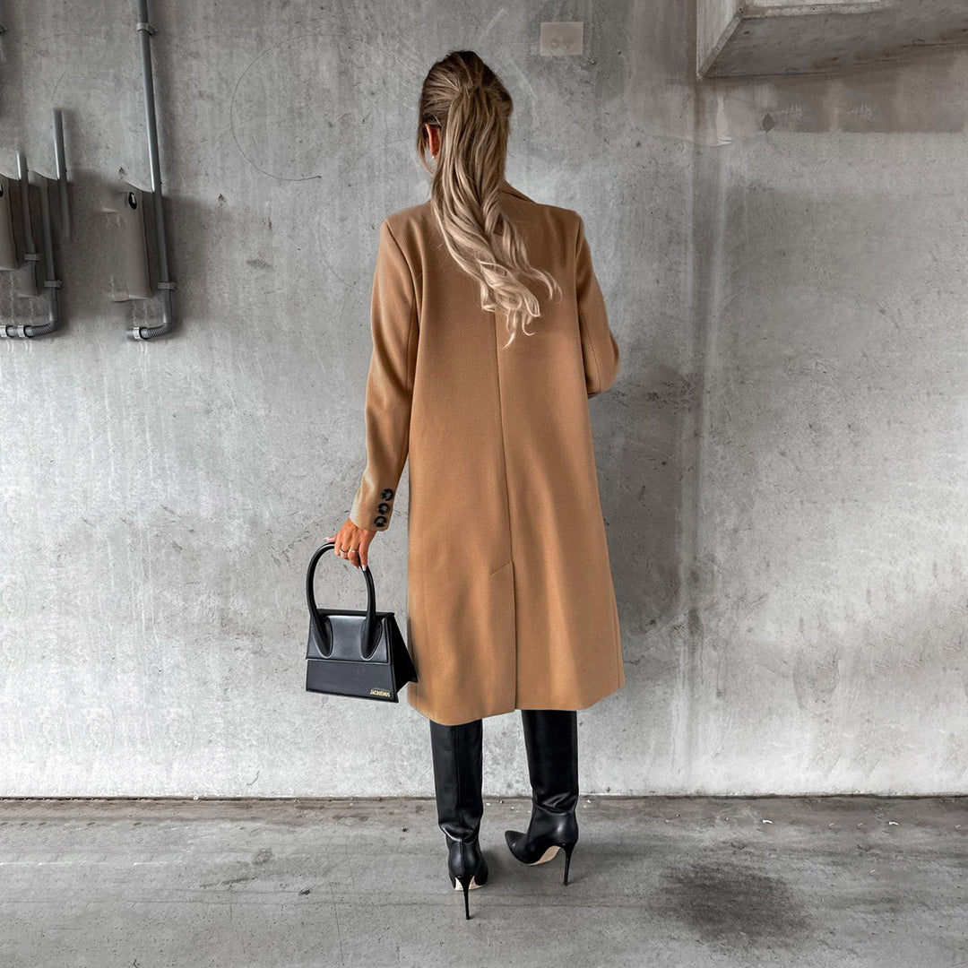 Frau in beigem Mantel, schwarze Stiefel, schwarze Handtasche, Betonhintergrund.