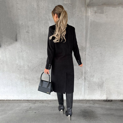 Frau in schwarzem Wintermantel, Lederstiefel, graue Handtasche, Betonhintergrund.