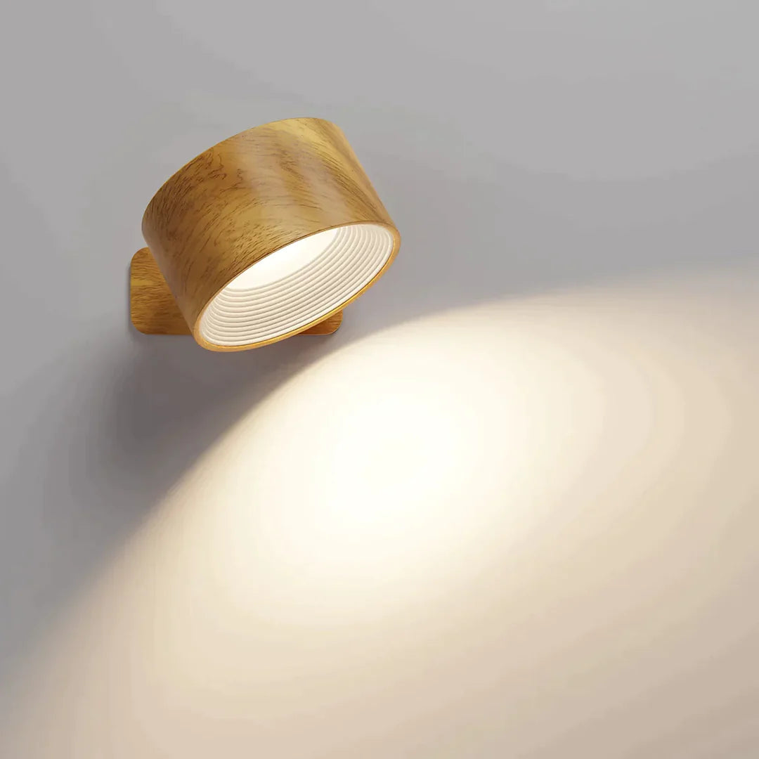 Vägglampa i träimitation med synlig ådring, tänd och riktad nedåt med en vit räfflad insida och varmt ljussken på väggen. 3