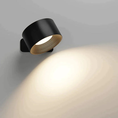 Svart vägglampa med guldfärgad räfflad insida, tänd och vinklad nedåt så att ljuset sprids i en mjuk båge på väggen. 2