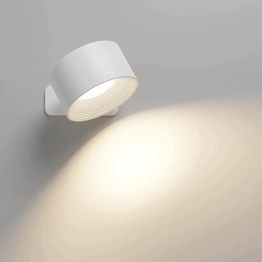 Vit cylinderformad vägglampa vinklad nedåt som kastar ett varmt ljuskägel över väggen, med räfflad insida kring ljuskällan. 1