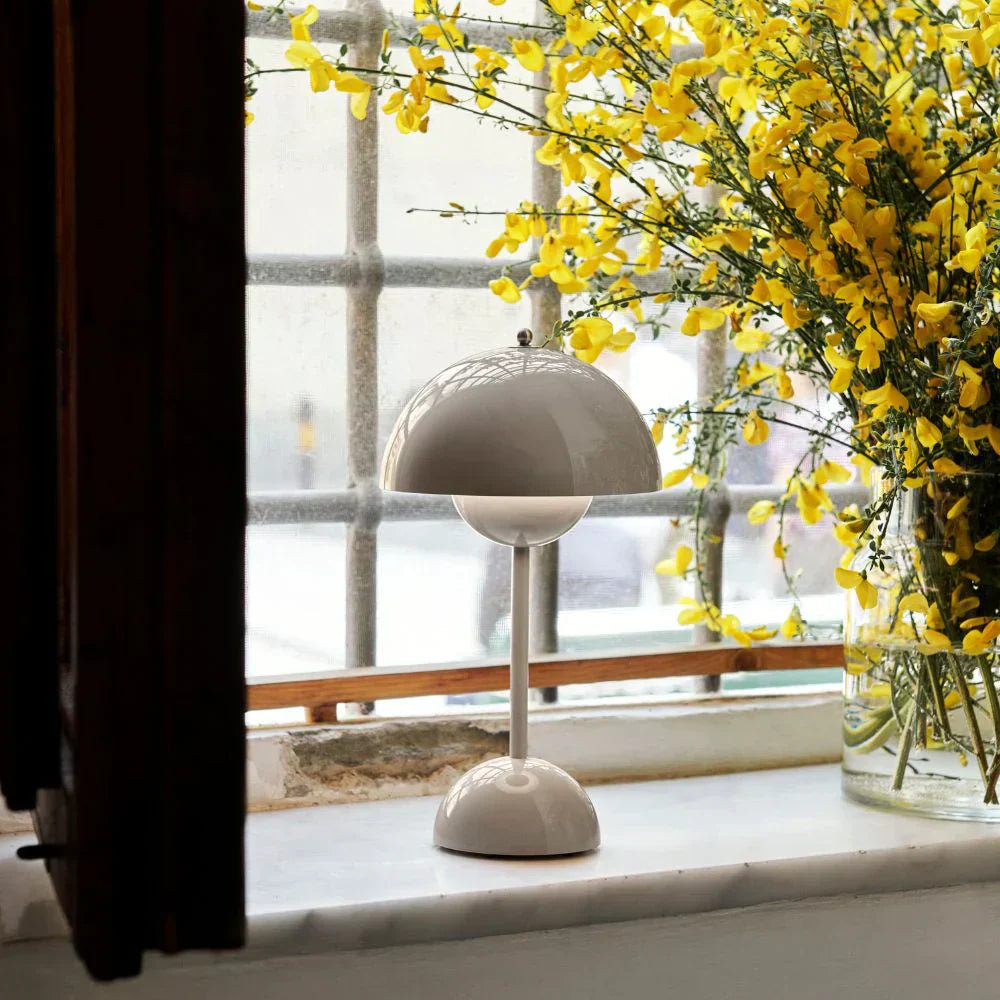 Graue Tischlampe auf Fensterbank, umgeben von gelben Blumen, modernes Design.
