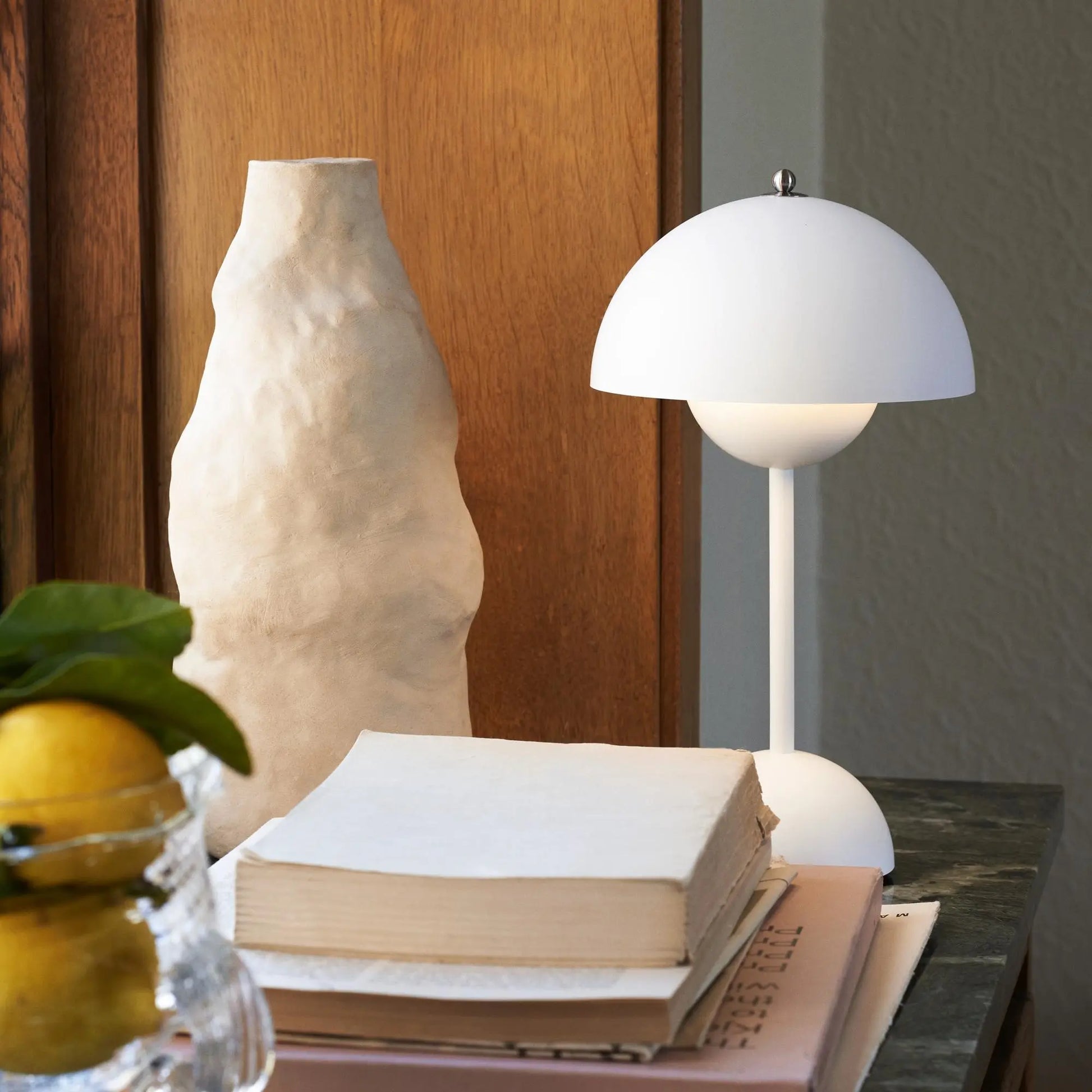 Weiße Tischlampe mit mattem Finish neben Keramikvase und Büchern auf Holzoberfläche.