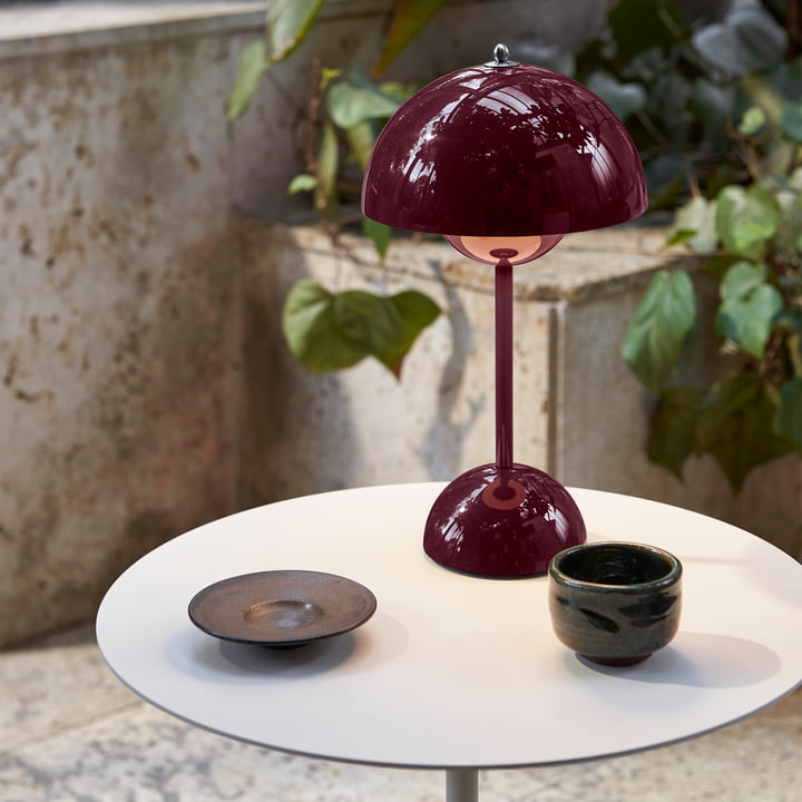 Weinrote Tischlampe aus Metall auf weißem Tisch, moderne Inneneinrichtung, Pflanzen im Hintergrund.