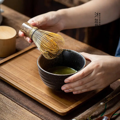 Hand hält Bambus-Teebesen über schwarzer Schale auf Holztablett, Matcha-Zubereitung, japanisch.