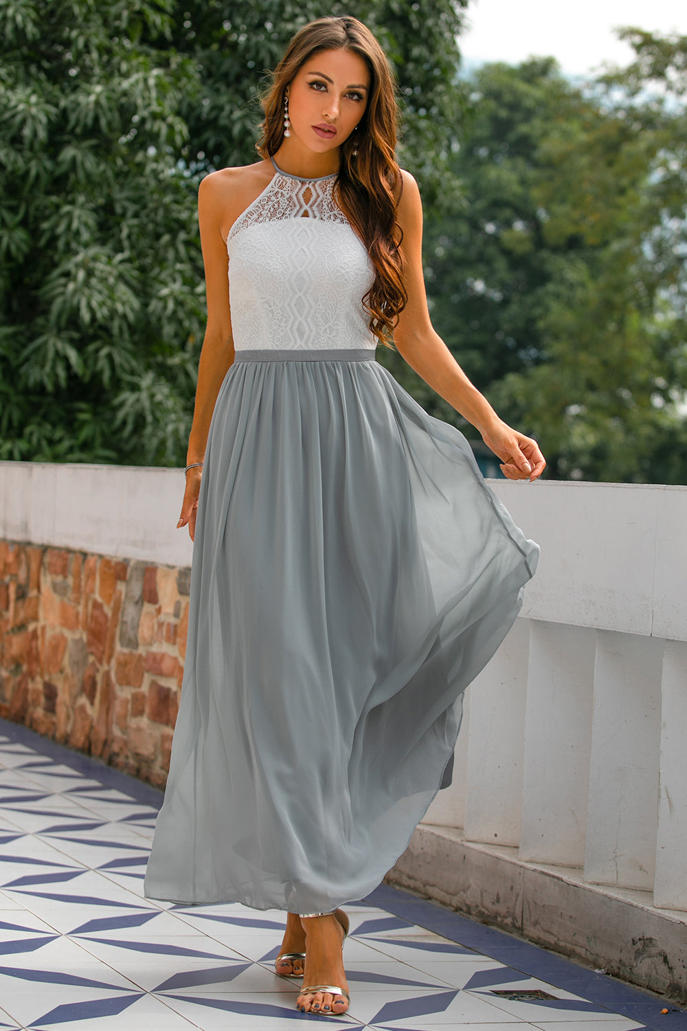 Frau im eleganten, ärmellosen Maxikleid aus Spitze und Chiffon in Weiß und Grau, draußen stehend.