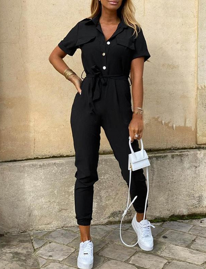 Frau in schwarzem Jumpsuit mit Gürtel, weiße Sneakers, kleine weiße Handtasche, modisch.