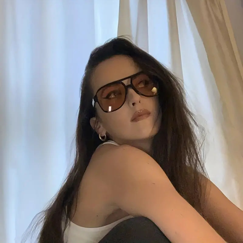 Frau mit brauner Sonnenbrille, weißem Top, langen Haaren vor hellem Vorhang.