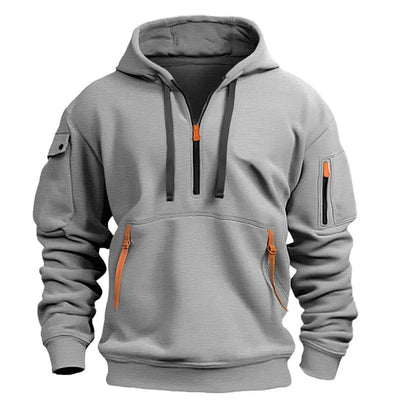 Ljusgrå hoodie sedd framifrån med halv dragkedja, svarta dragsnören, orange dragkedjesliders och dragkedjefickor på kängurufickan och ärmen. 1