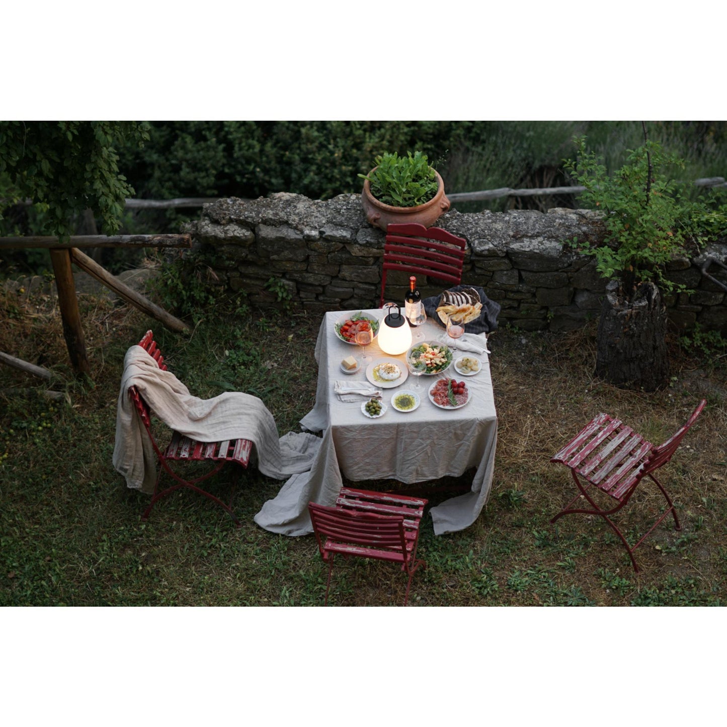 Gedeckter Gartentisch mit Speisen, roter Stuhl, Steinmauer, Abendessen im Freien, stimmungsvolle Beleuchtung.