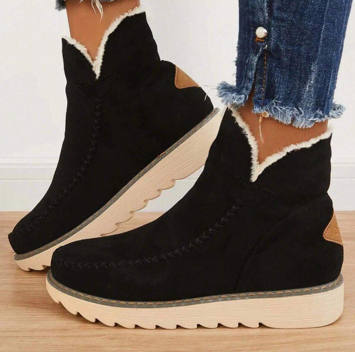 Svarta mockaliknande ankelboots med vit fleecekant och beige räfflad sula, sedda från sidan på en modell i fransiga jeans. 1