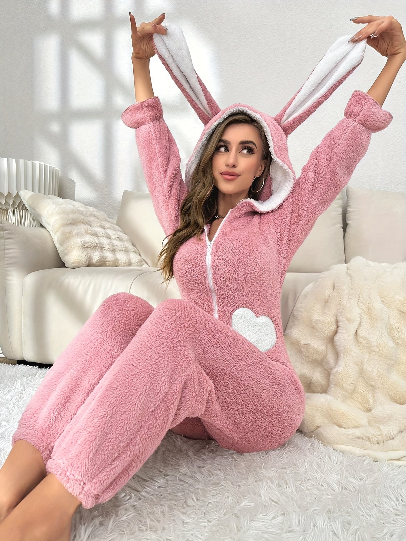 Frau im rosa Hasen-Overall mit Kapuze, Herzmotiv, auf weißem Sofa, gemütliche Wohnzimmeratmosphäre.