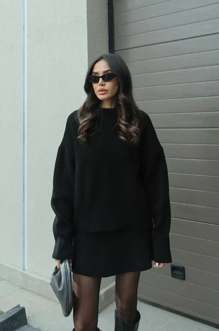 Modell utomhus i svart stickad uppsättning med oversize-tröja och kort kjol, med tunna strumpbyxor, höga skor och grå väska. 5