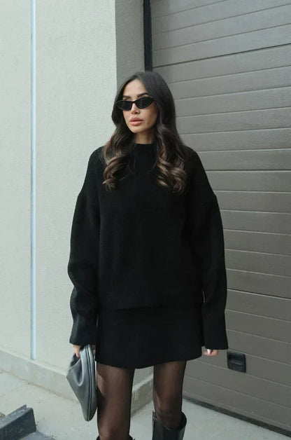 Modell utomhus i svart stickad uppsättning med oversize-tröja och kort kjol, med tunna strumpbyxor, höga skor och grå väska. 5