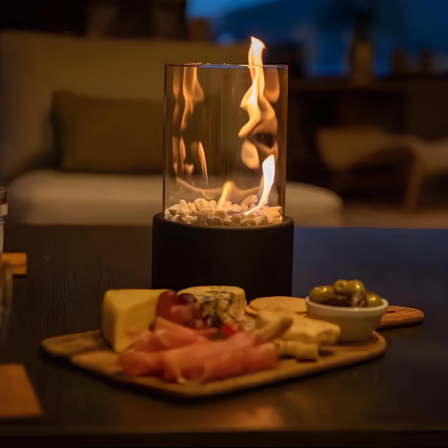 Tischkamin mit Flamme, umgeben von Käseplatte und Oliven, gemütliche Wohnzimmeratmosphäre.