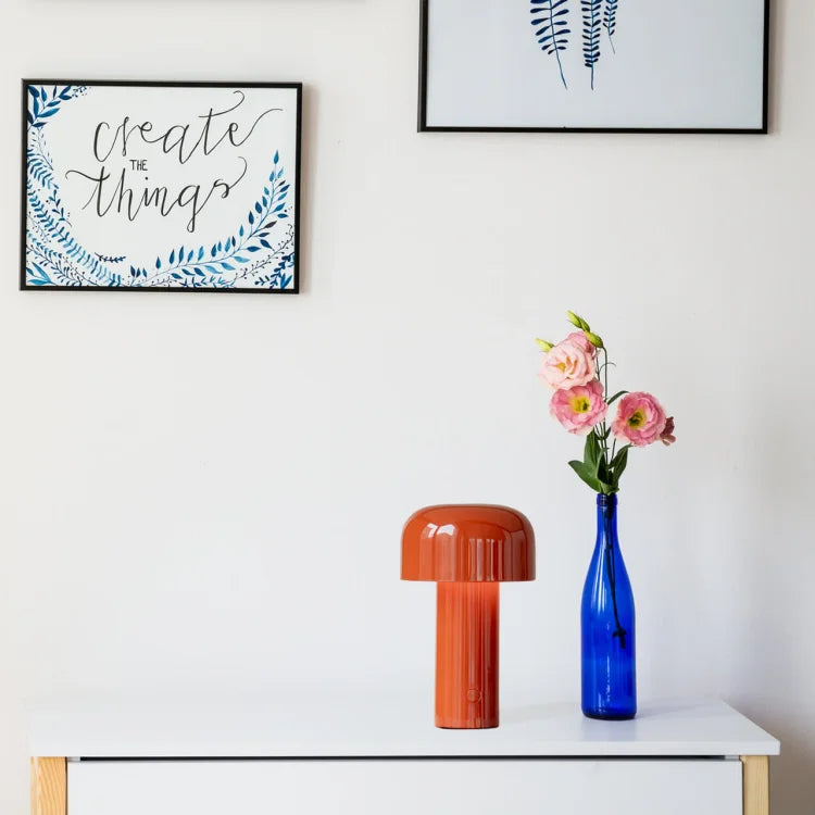 Orangefarbene Tischlampe aus Keramik auf weißem Tisch, blaue Vase mit rosa Blumen daneben.