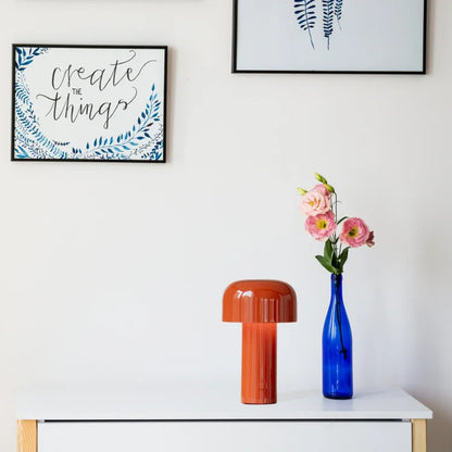 Orangefarbene Tischlampe aus Keramik auf weißem Tisch, blaue Vase mit rosa Blumen daneben.