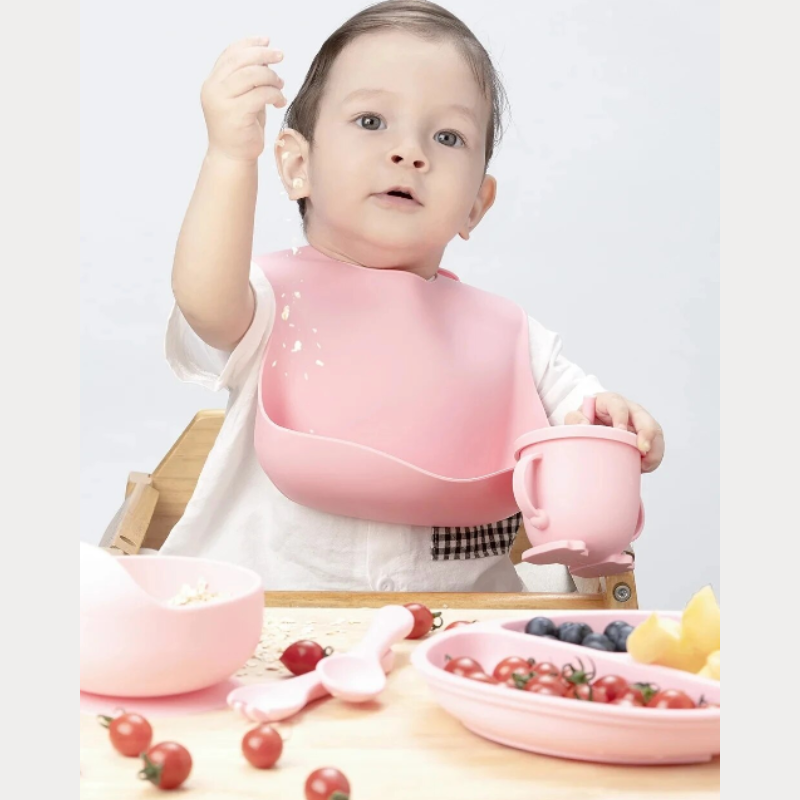 Kleinkind mit rosa Lätzchen isst am Tisch, umgeben von Geschirr und frischem Obst.