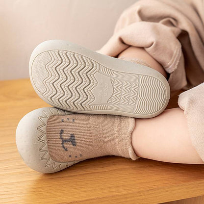 Kleinkind trägt beige rutschfeste Hausschuhe mit Gummisohle auf Holzoberfläche.
