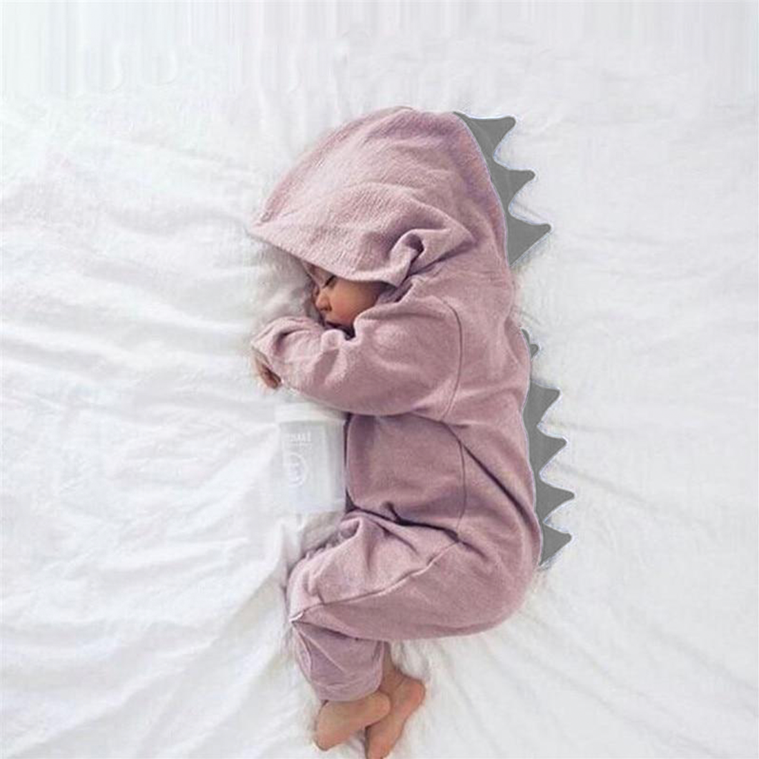 Schlafender Säugling im rosa Dinosaurier-Strampler mit Kapuze auf weißem Bett.