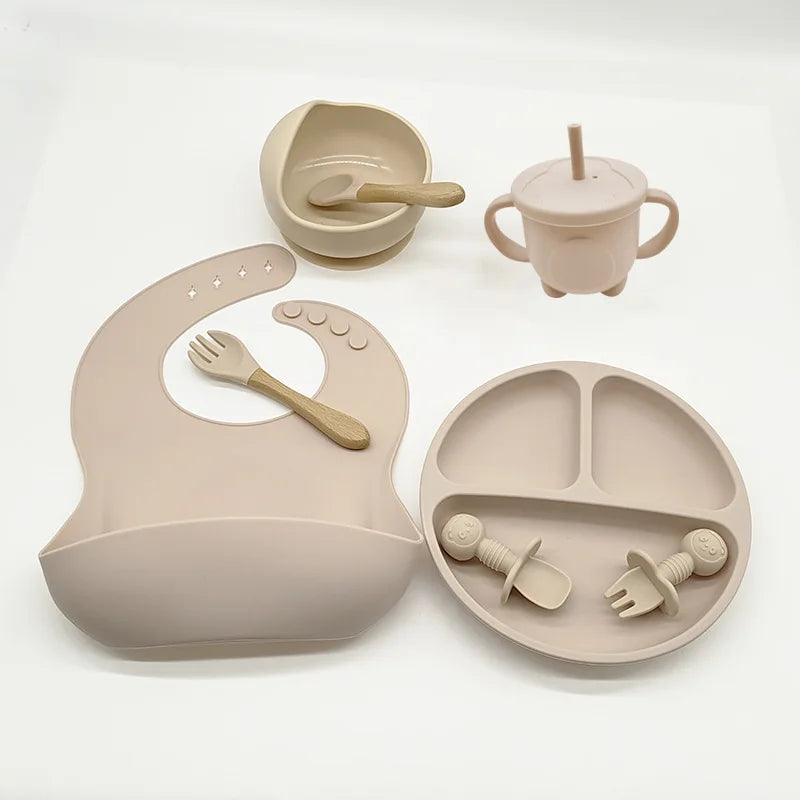 Beiges Babygeschirr-Set aus Silikon, bestehend aus Lätzchen, Teller, Becher und Besteck.