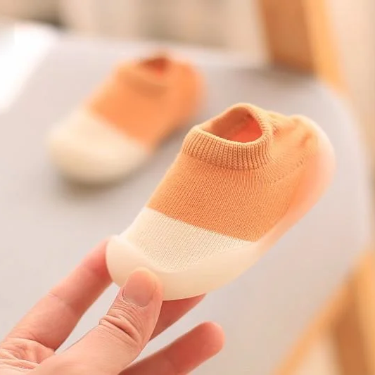 Orange-weiße Babyschuhe aus Baumwolle, rutschfest, handgehalten, auf grauem Hintergrund.