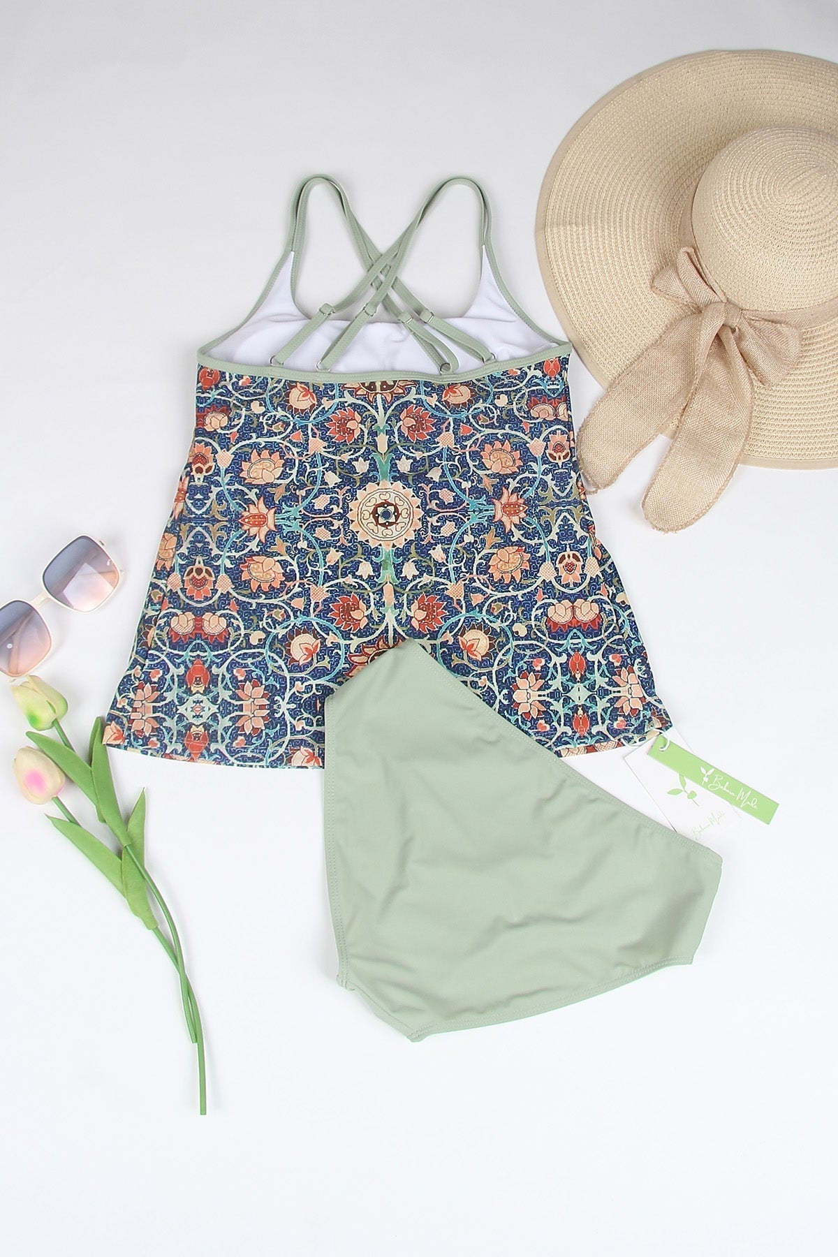 Badeanzug mit floralem Muster, grünem Rock, Strohhut, Sonnenbrille und Tulpe auf weißem Hintergrund.