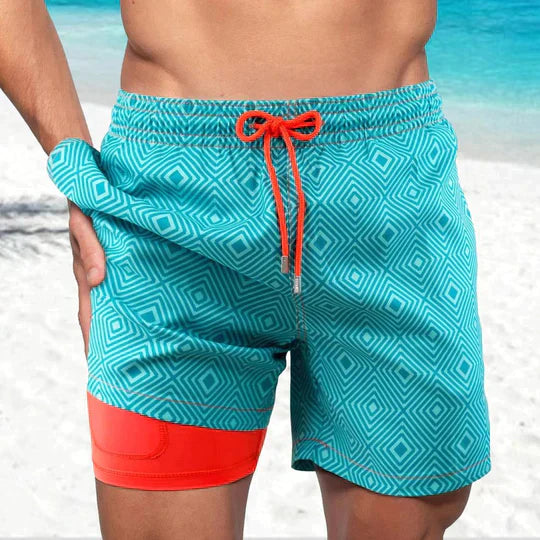 Herren-Badeshorts türkis mit geometrischem Muster und orangefarbenem Bund am Strand.