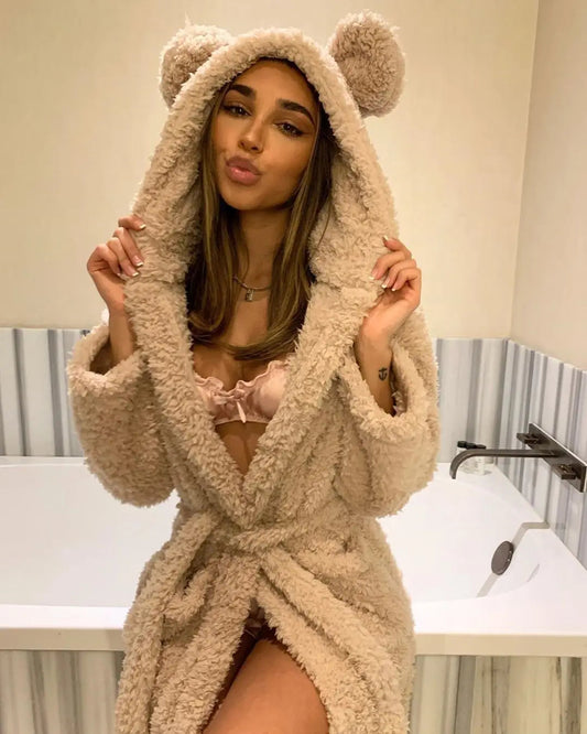 Frau in beigem Teddyfleece-Bademantel mit Kapuze und Ohren, sitzt vor Badewanne.