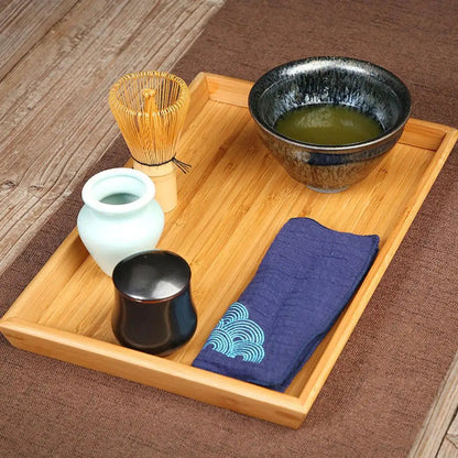 Bambus-Teeset mit Matcha-Schale, Bambusbesen, Keramikgefäß und blauem Tuch auf Holztablett.