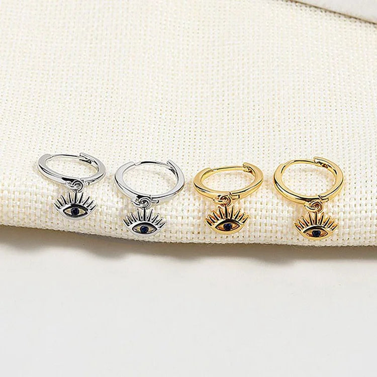 Silberne und goldene Ohrringe mit Auge-Anhänger auf beigem Stoff, modischer Schmuck.