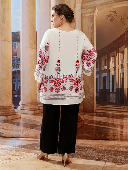 Frau in weißer Tunika mit roten Blumenmustern, schwarze Hose, elegante Umgebung.