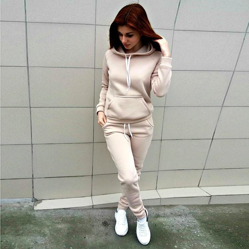 Frau in beigem Hoodie-Trainingsanzug, Kapuze, Kängurutasche, weiße Sneaker, moderne Sportmode.